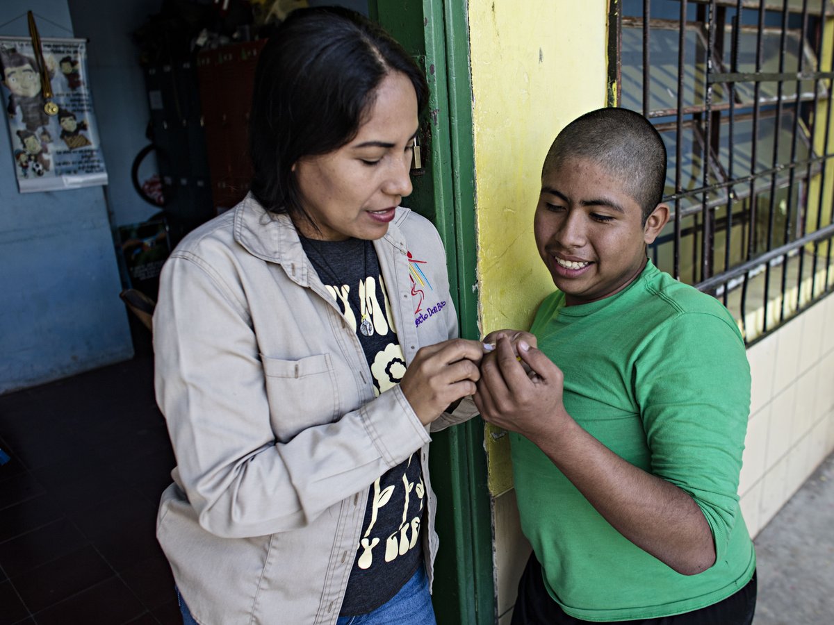 Eine Frau und ein Junge tauschen lächelnd etwas Kleines in einer Türöffnung aus, im Hintergrund eine gelbe Wand mit Gitterfenster.