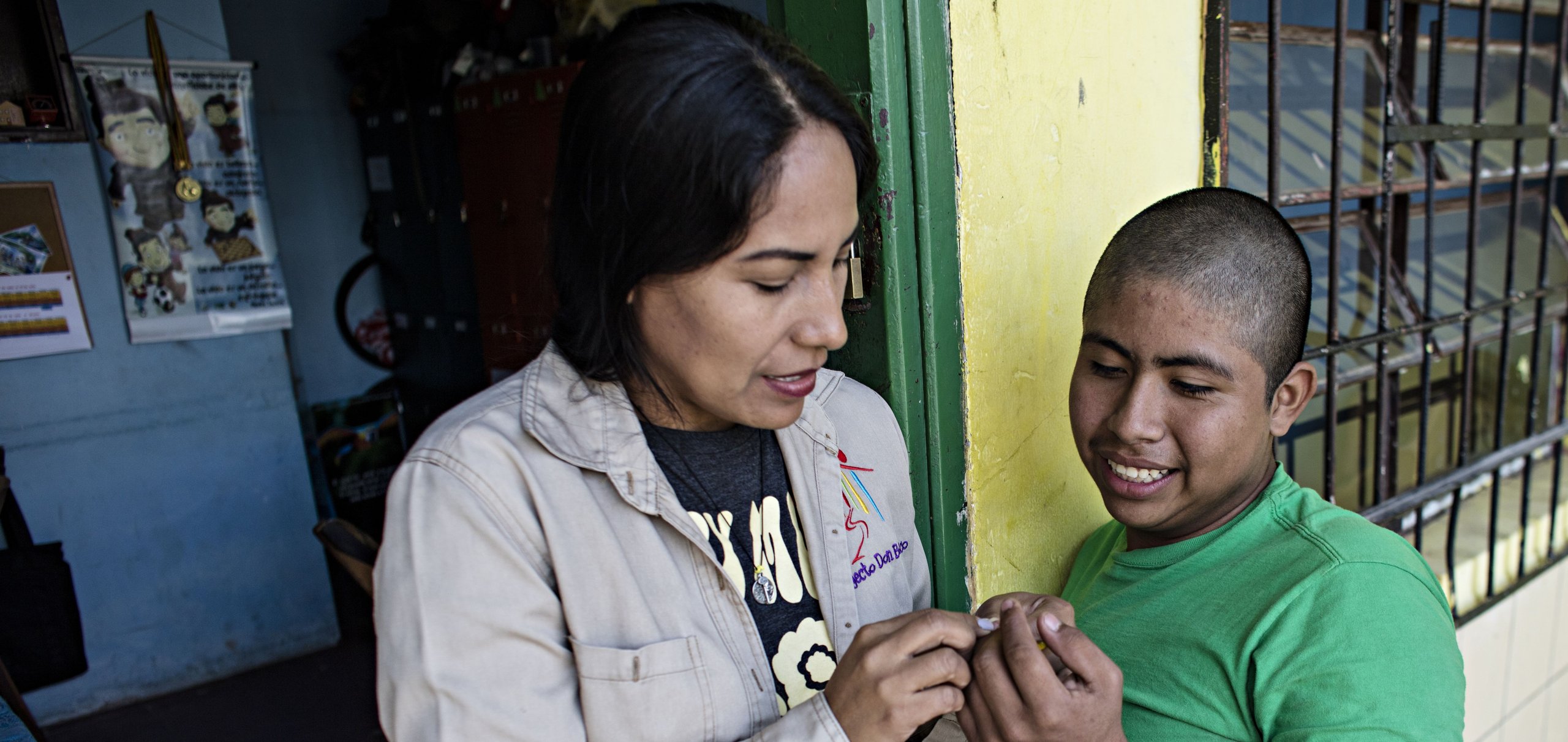 Eine Frau und ein Junge tauschen lächelnd etwas Kleines in einer Türöffnung aus, im Hintergrund eine gelbe Wand mit Gitterfenster.