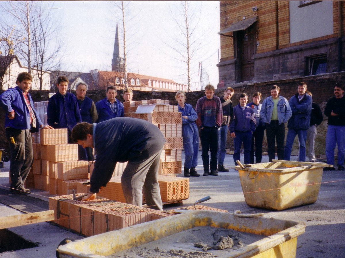 Bauarbeiter, meist Männer, beobachten einen Kollegen beim Mauern auf einer Baustelle mit Ziegelstapeln und Mörtelwannen.