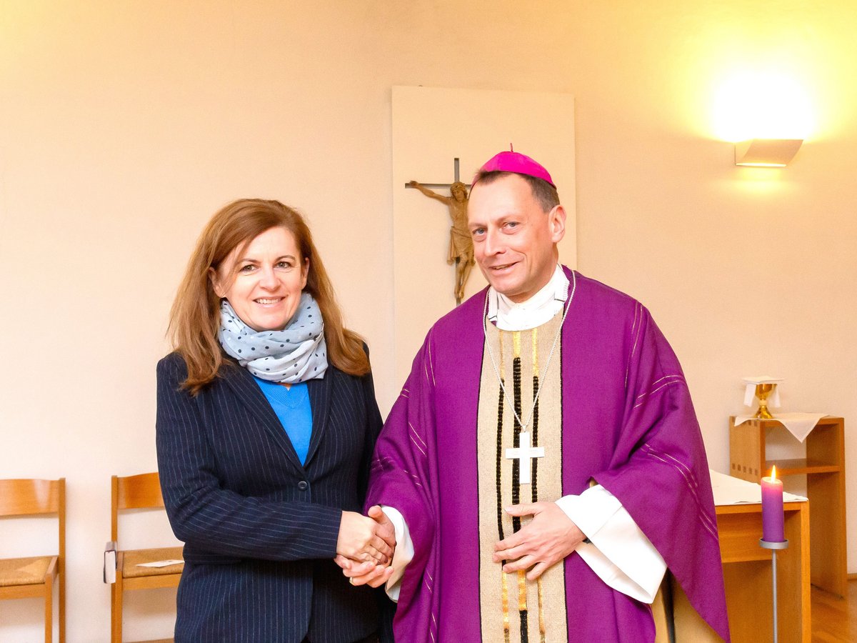 Eine lächelnde Frau in gestreifter Jacke schüttelt einem lächelnden Bischof in lila Gewändern die Hand, Kruzifix im Hintergrund.