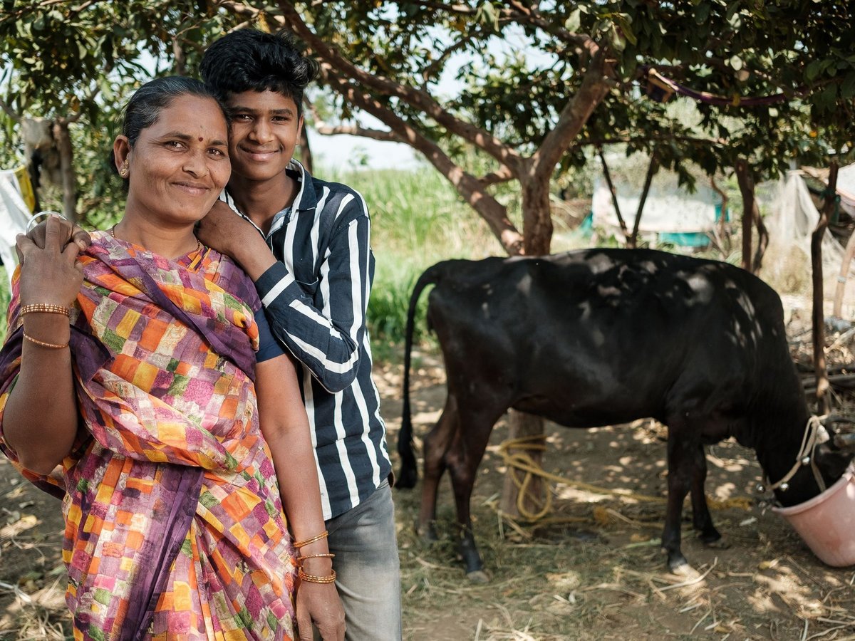 Eine lächelnde Frau im bunten Sari umarmt einen lächelnden jungen Mann. Im Hintergrund ist eine Kuh mit einem Eimer zu sehen.