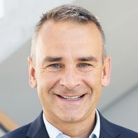 Lächelnder Mann mit kurzen grauen Haaren und blauen Augen in dunklem Anzug und hellblauem Hemd.