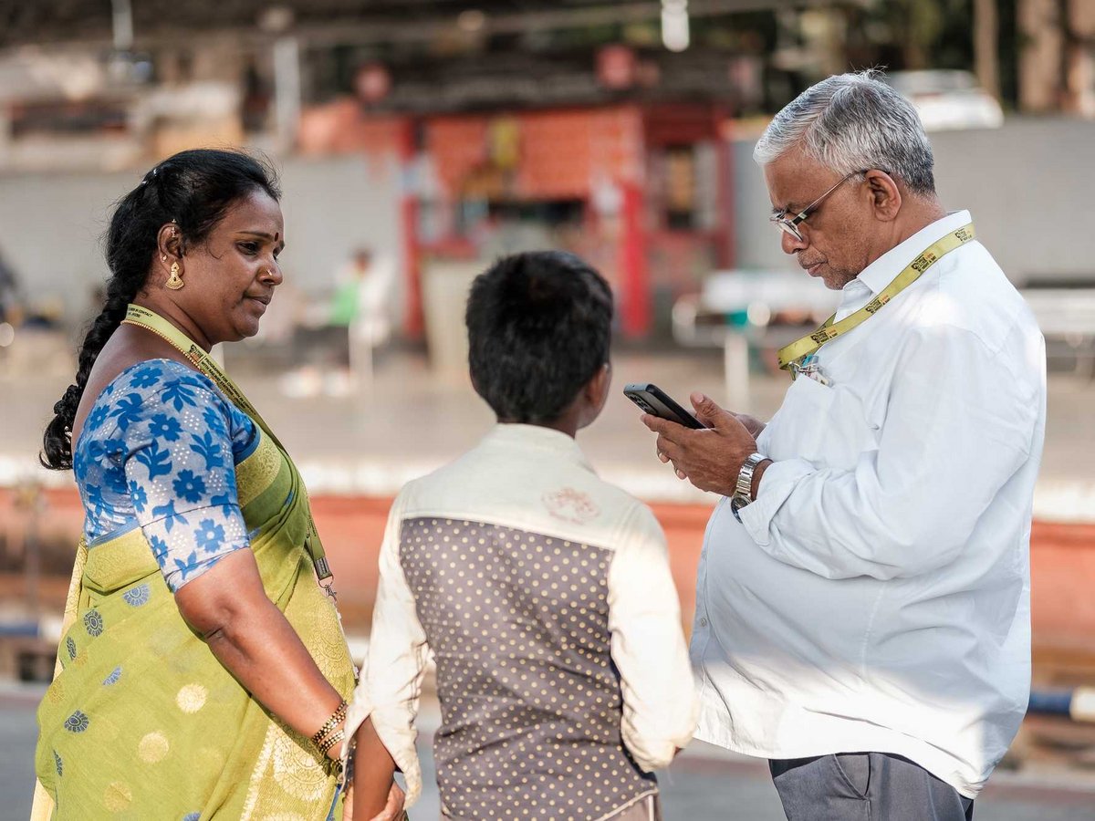 Eine Frau im Sari, ein Kind und ein Mann, der auf sein Smartphone schaut, an einem öffentlichen Ort in Indien.