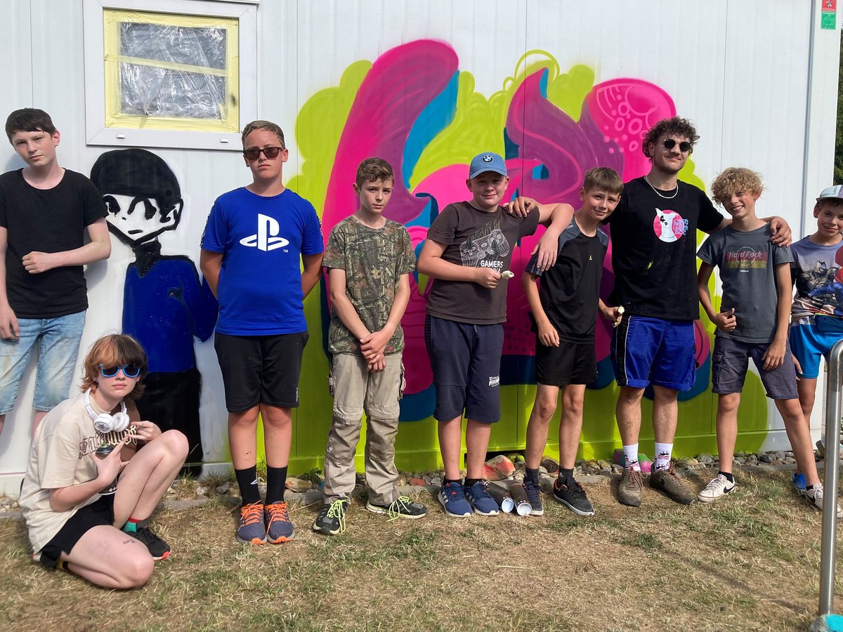 Zehn Jungen und junge Männer vor einer weißen Wand mit bunten Graffiti. Ein Junge kniet am Boden.