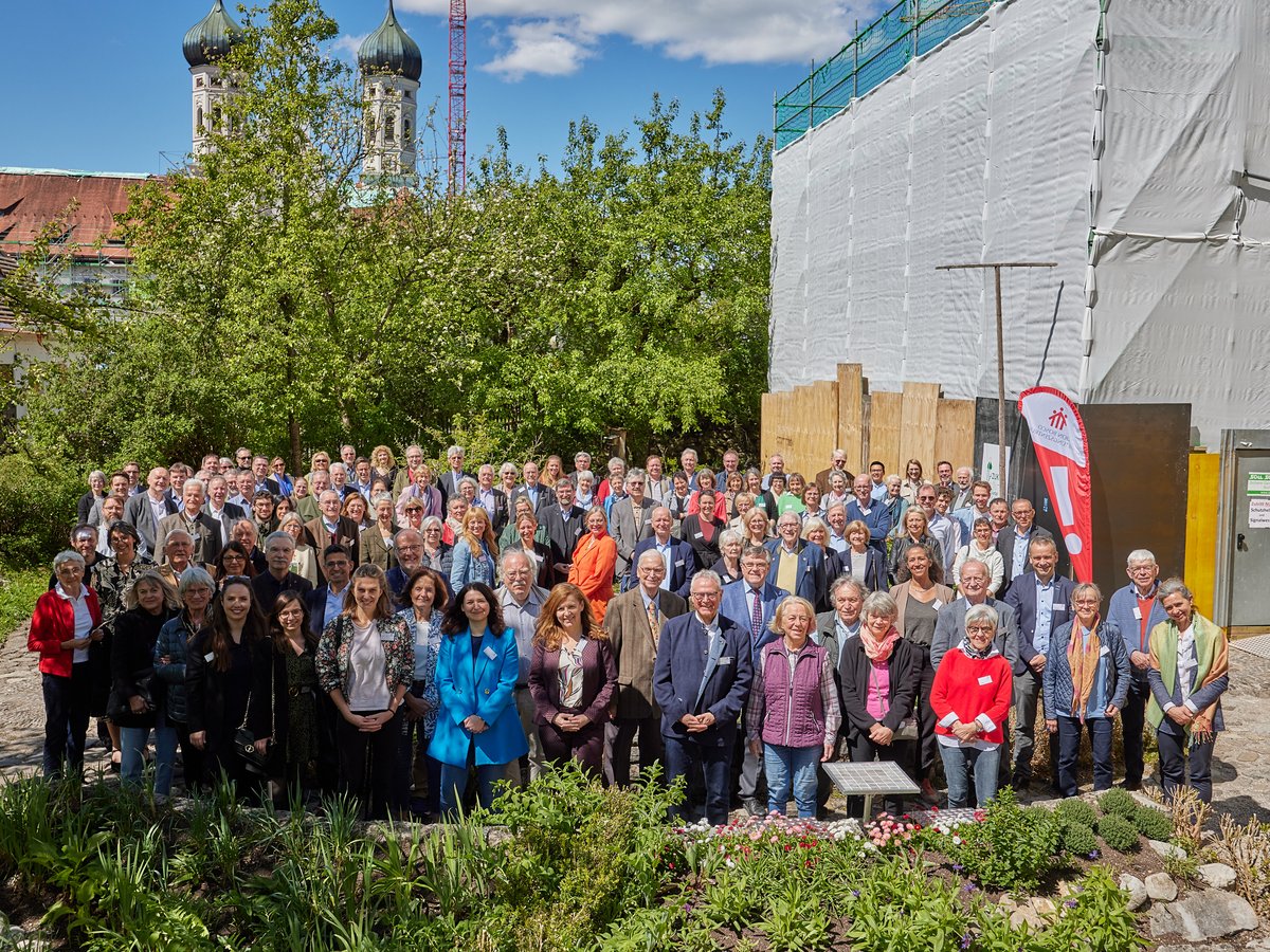 Große Gruppe lächelnder Menschen im Freien vor einer Kirche, Bäumen und einer Baustelle unter blauem Himmel an einem sonnigen Tag.