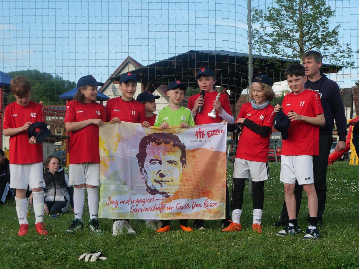 Eine Gruppe von Kindern in Sportkleidung hält ein Don Bosco-Banner, einer hält einen Pokal und ein Mikrofon. Ein Erwachsener steht daneben.