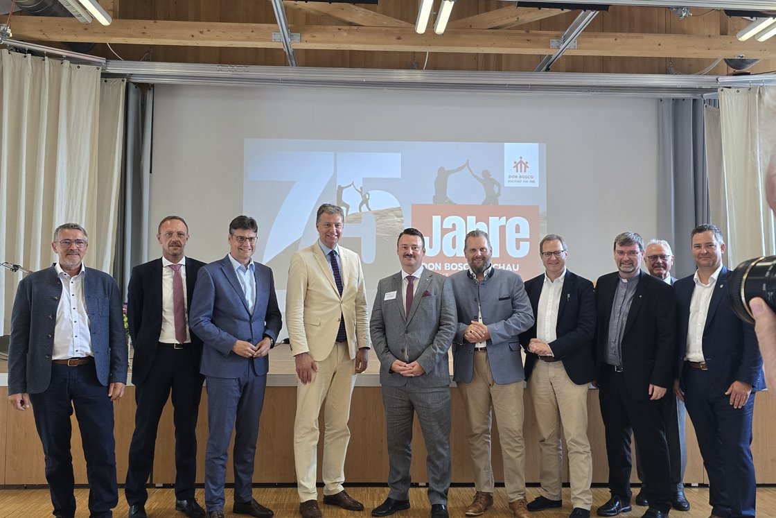 Gruppe von elf Männern in Anzügen und traditioneller Kleidung posiert vor einem Bildschirm mit '75 Jahre' Logo.