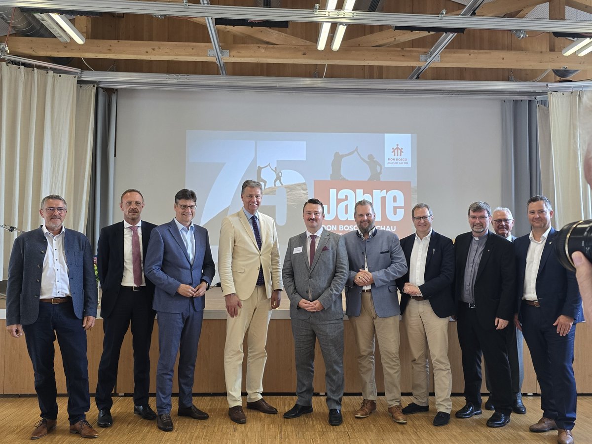 Gruppe von elf Männern in Anzügen und traditioneller Kleidung posiert vor einem Bildschirm mit '75 Jahre' Logo.