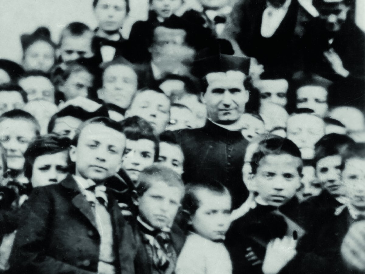 Schwarz-Weiß-Foto eines Priesters in Soutane, umgeben von vielen kleinen Jungen, alle blicken in die Kamera.