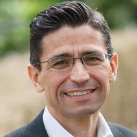 Porträt eines lächelnden Mannes mit dunklem Haar und Brille, der ein weißes Hemd und eine dunkle Jacke trägt, vor verschwommenem grünem Hintergrund.