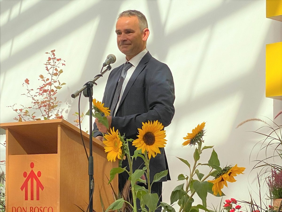 Ein Mann im dunklen Anzug steht hinter einem Holzpult mit Don-Bosco-Logo und hält eine Sonnenblume. Sonnenblumen und Pflanzen schmücken die Szene.
