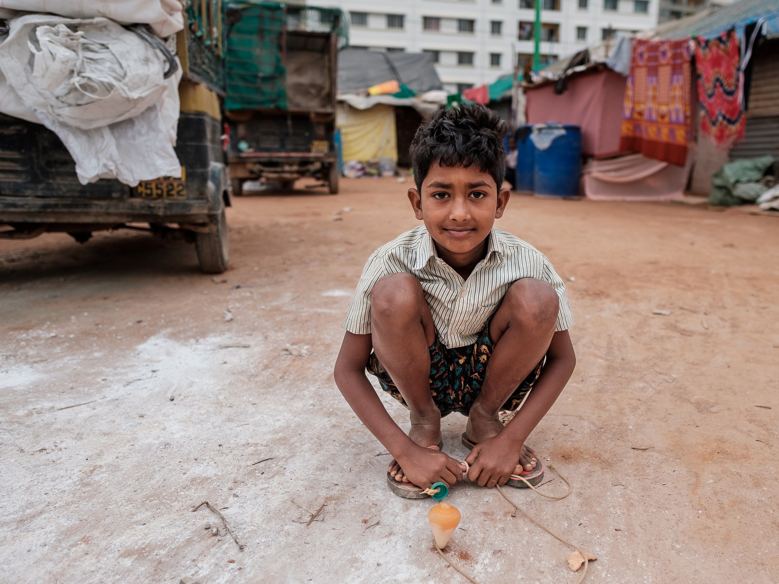 Ein junger Junge hockt lächelnd auf staubigem Boden, hält einen Kreisel mit Schnur, im Hintergrund provisorische Behausungen.