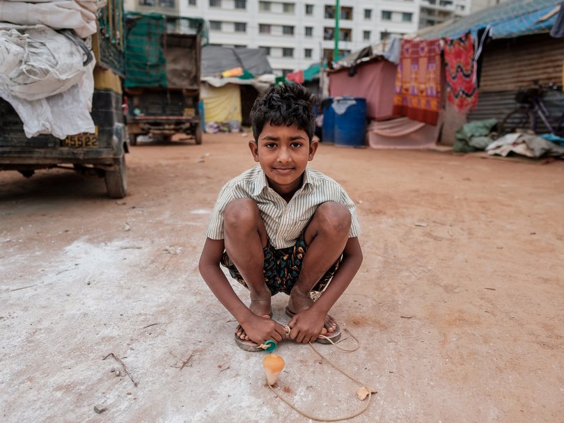 Ein junger Junge hockt lächelnd auf staubigem Boden, hält einen Kreisel mit Schnur, im Hintergrund provisorische Behausungen.