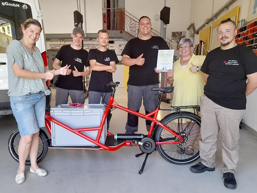 Gruppe von sechs lächelnden Personen mit Daumen hoch neben einem roten Lastenrad, eine Urkunde haltend in einer Werkstatt.