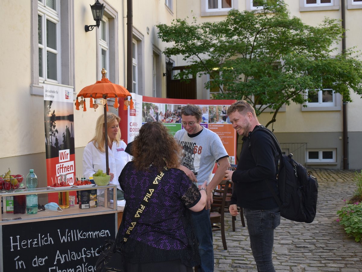 Personen an einem Outdoor-Infostand der 'Anlaufstelle für Ehemalige' mit Kreidetafel 'Herzlich Willkommen', Obst und Schirm.