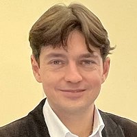 Kopfaufnahme eines Mannes mit dunklem, gewelltem Haar, leicht lächelnd, in weißem Hemd und dunklem Sakko vor hellem Hintergrund.