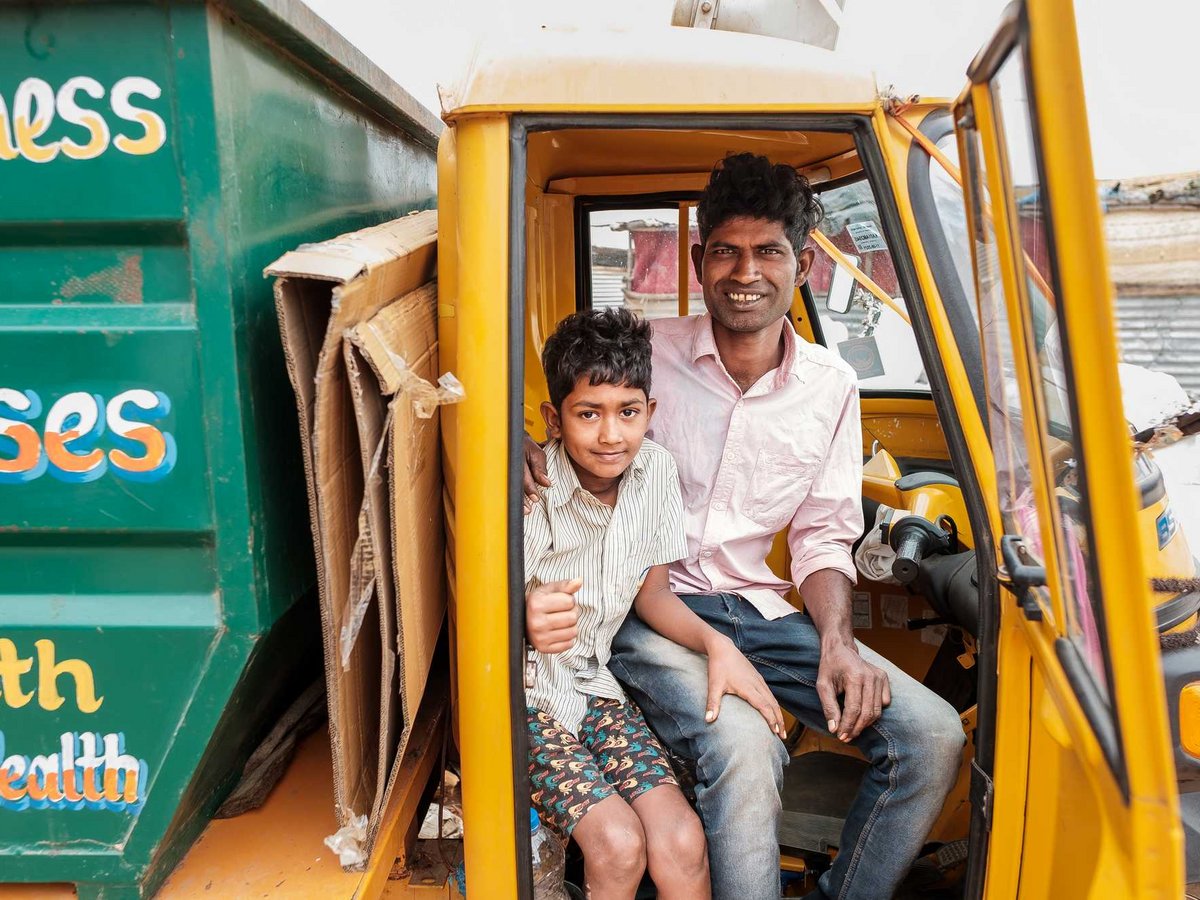 Ein lächelnder Mann und Junge sitzen in einer gelben Autorikscha neben Pappe und einem grünen Behälter mit der Aufschrift 'Health'.