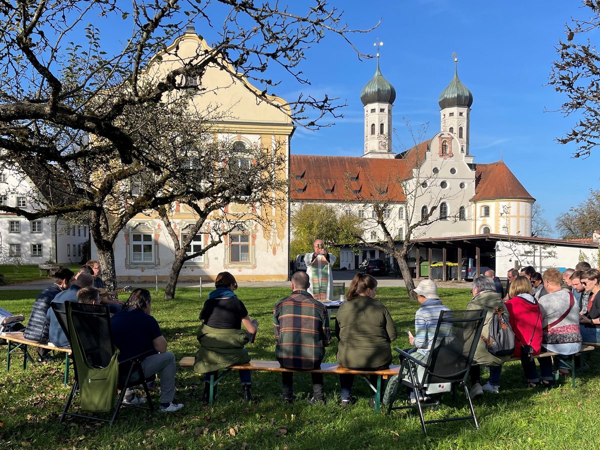 Open-Air-Gottesdienst mit Priester und Gläubigen vor historischen Barockgebäuden mit Zwiebeltürmen bei Sonnenschein.
