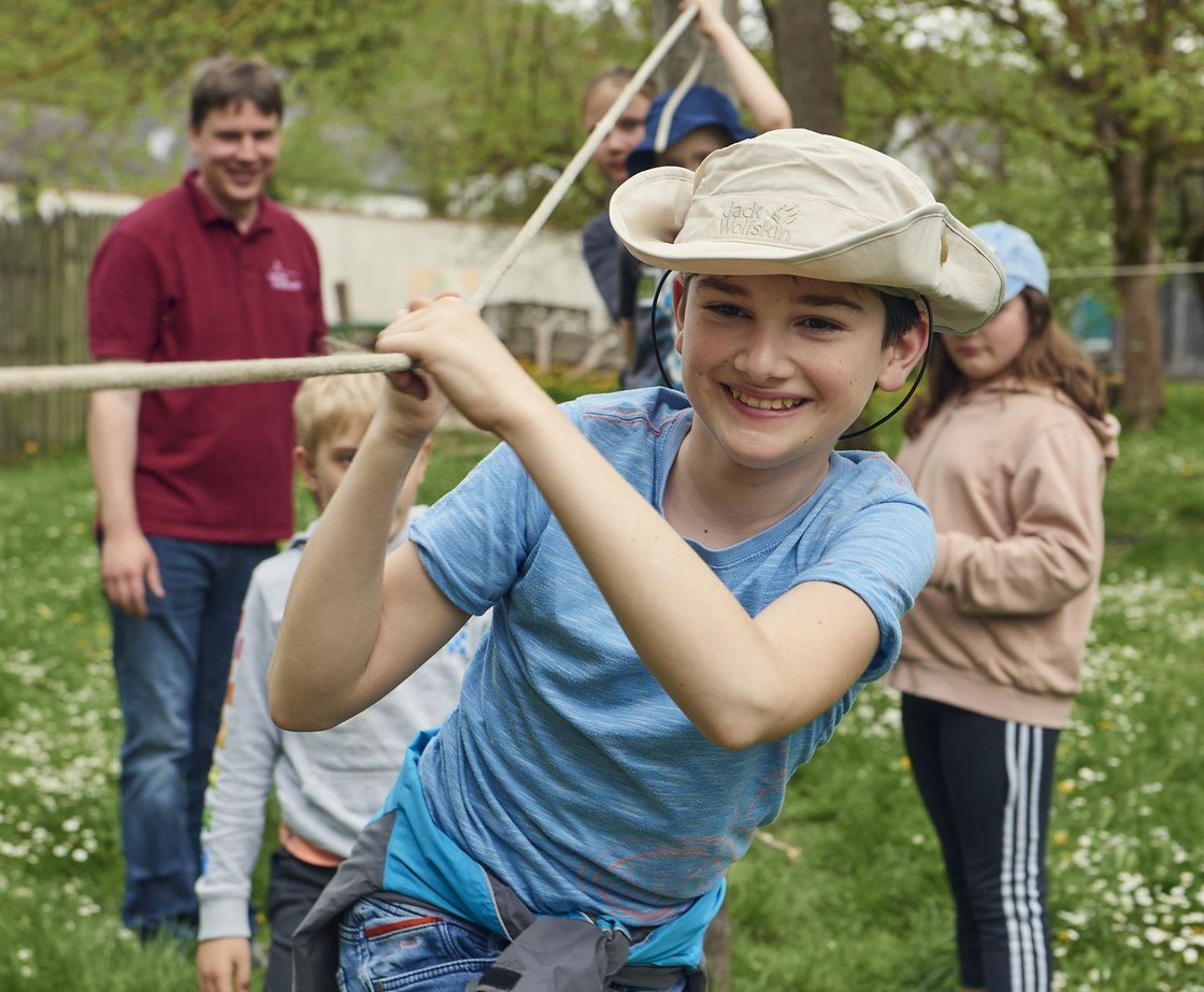 Lächelnder Junge mit Hut hält ein Seil bei einer Outdoor-Gruppenaktivität mit anderen Kindern und einem Erwachsenen.