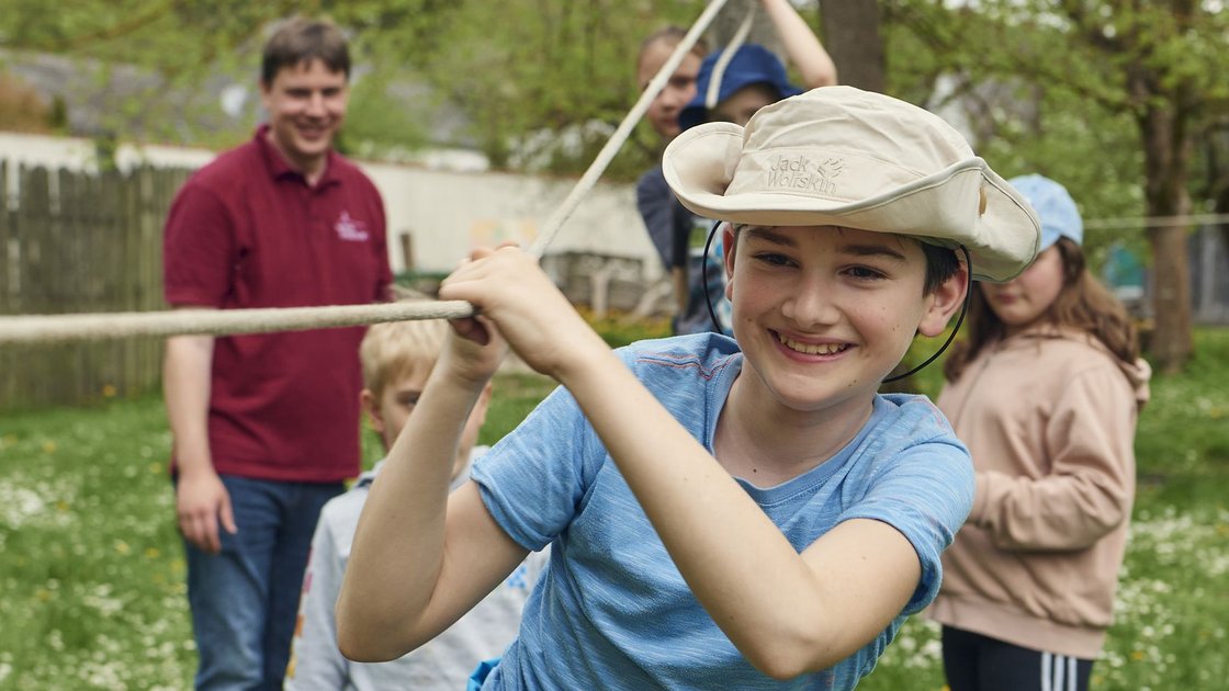 Lächelnder Junge mit Hut hält ein Seil bei einer Outdoor-Gruppenaktivität mit anderen Kindern und einem Erwachsenen.