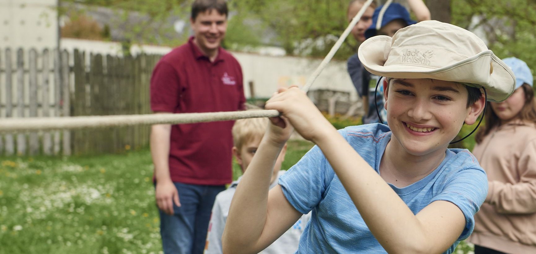 Lächelnder Junge mit Hut hält ein Seil bei einer Outdoor-Gruppenaktivität mit anderen Kindern und einem Erwachsenen.