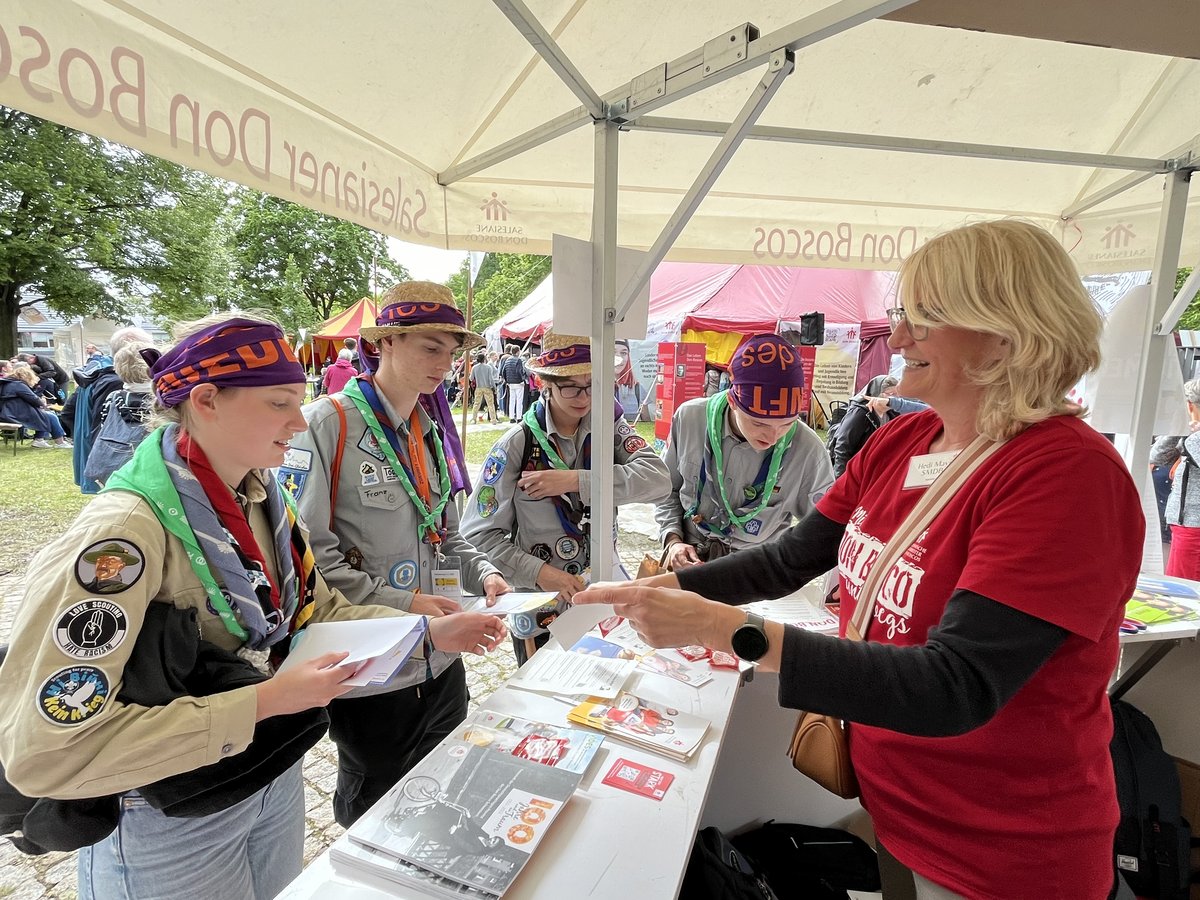 Eine lächelnde Frau überreicht drei Pfadfindern Informationsmaterial an einem Don Bosco Stand unter einem Zelt.