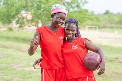 Zwei lächelnde junge Frauen in Don Bosco Sportuniformen auf einem Feld, eine zeigt Daumen hoch, die andere hält einen Ball.