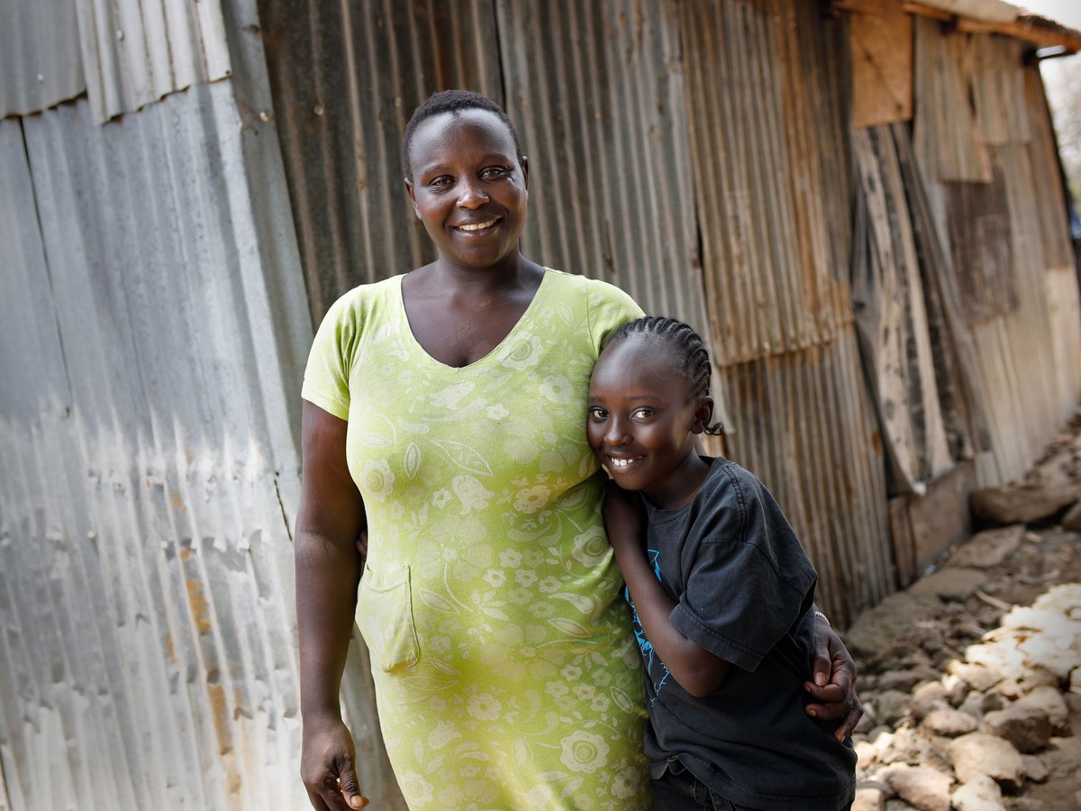 Lächelnde afrikanische Frau und Kind umarmen sich vor einer Wellblechwand.