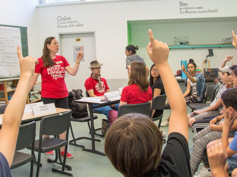Eine Lehrerin gestikuliert während sie Schüler in einem Klassenzimmer unterrichtet, viele mit erhobenen Händen bei einer Lektion über Solidarität.