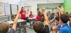 Eine Lehrerin gestikuliert während sie Schüler in einem Klassenzimmer unterrichtet, viele mit erhobenen Händen bei einer Lektion über Solidarität.