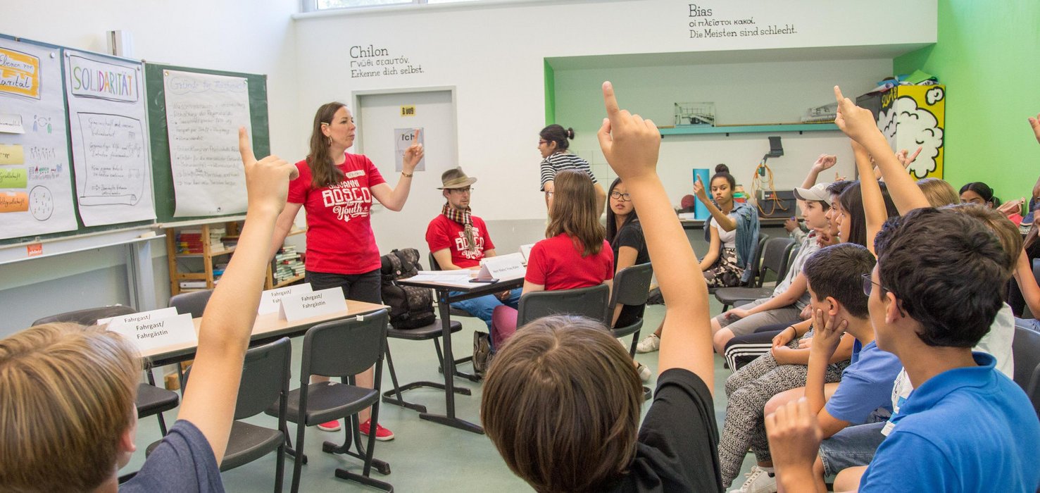 Eine Lehrerin gestikuliert während sie Schüler in einem Klassenzimmer unterrichtet, viele mit erhobenen Händen bei einer Lektion über Solidarität.