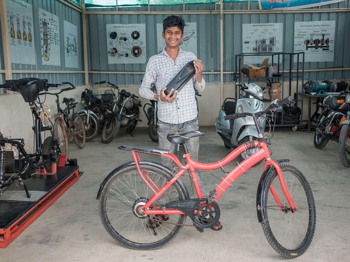 Lächelnder junger Mann hält ein E-Bike-Bauteil neben einem roten Elektrofahrrad in einer Werkstatt voller Fahrzeuge.