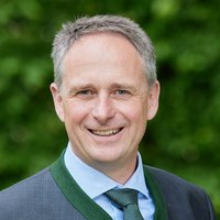 Lächelnder Mann mit grauen Haaren, blauem Hemd und grün gemusterter Krawatte, trägt einen Trachtenjanker vor grünem Hintergrund.