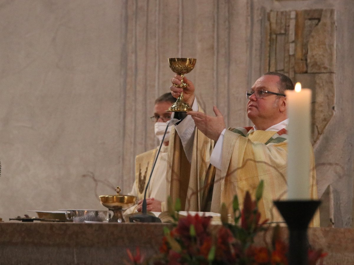 Ein Priester erhebt einen goldenen Kelch während einer religiösen Zeremonie. Monstranz und Kerze auf dem Altar sichtbar.