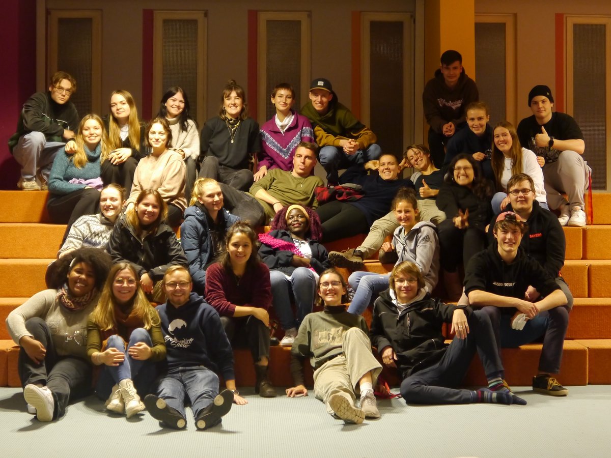 Eine große Gruppe lächelnder junger Erwachsener sitzt auf orangefarbenen Stufen und dem Boden und blickt in die Kamera.