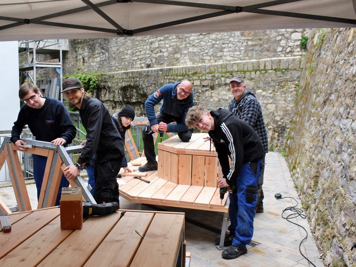 Eine Gruppe von Personen baut gemeinsam eine Holzkonstruktion im Freien, einige lächeln in die Kamera.