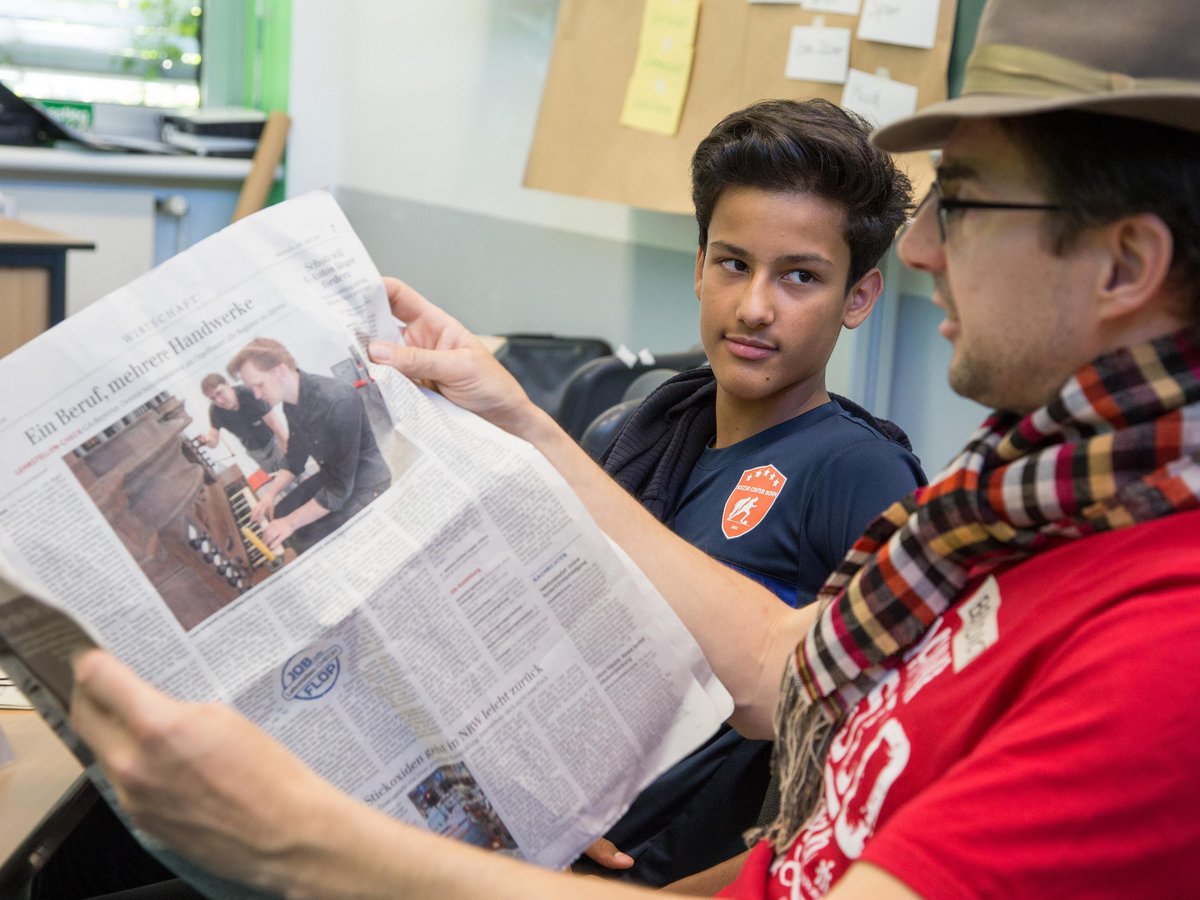 Ein Mann mit Hut liest einem jungen Mann in blauem T-Shirt eine Zeitung vor, der interessiert auf einen Artikel über Handwerke blickt.