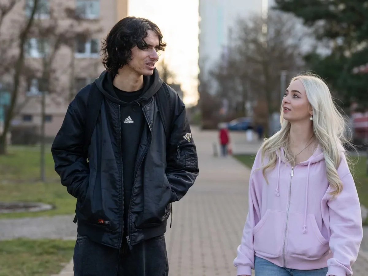 Ein Mann mit dunklen Haaren und eine blonde Frau in rosa Hoodie spazieren im Freien und unterhalten sich.