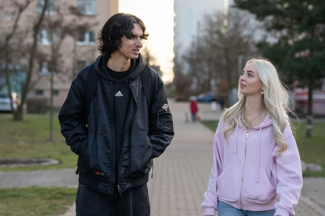 Ein Mann mit dunklen Haaren und eine blonde Frau in rosa Hoodie spazieren im Freien und unterhalten sich.