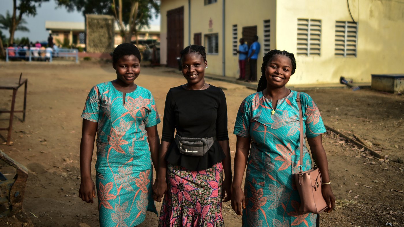 Drei junge Frauen lächeln in die Kamera, zwei tragen passende Kleider, die dritte eine Bluse und Rock, vor einem Gebäude.