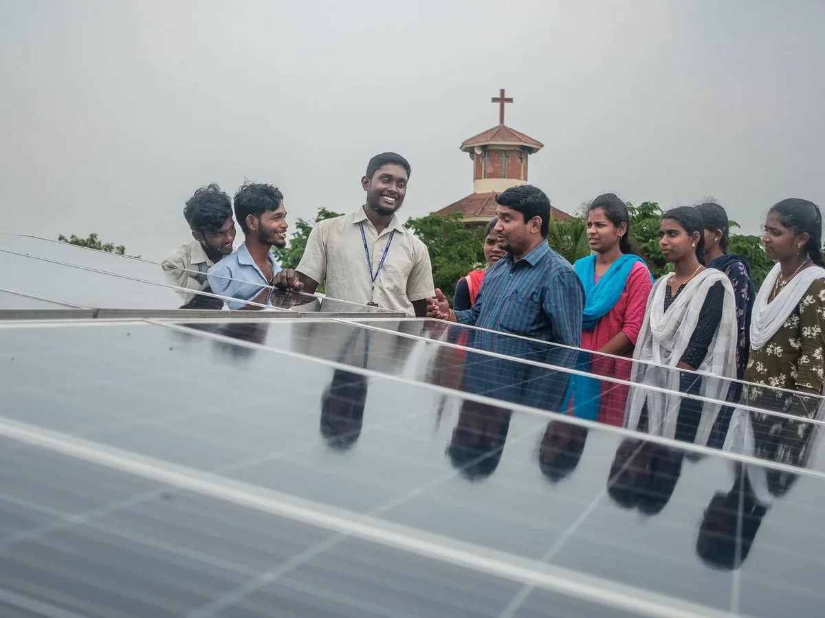 Gruppe von Schülern und Lehrern lernt auf einem Dach über Solarpaneele, mit einem Kirchengebäude im Hintergrund.