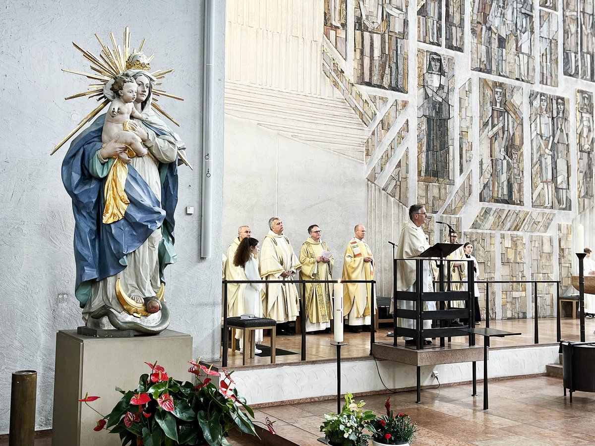 Marienstatue und Priester bei einem Gottesdienst in einer modernen Kirche mit großer Mosaikwand.