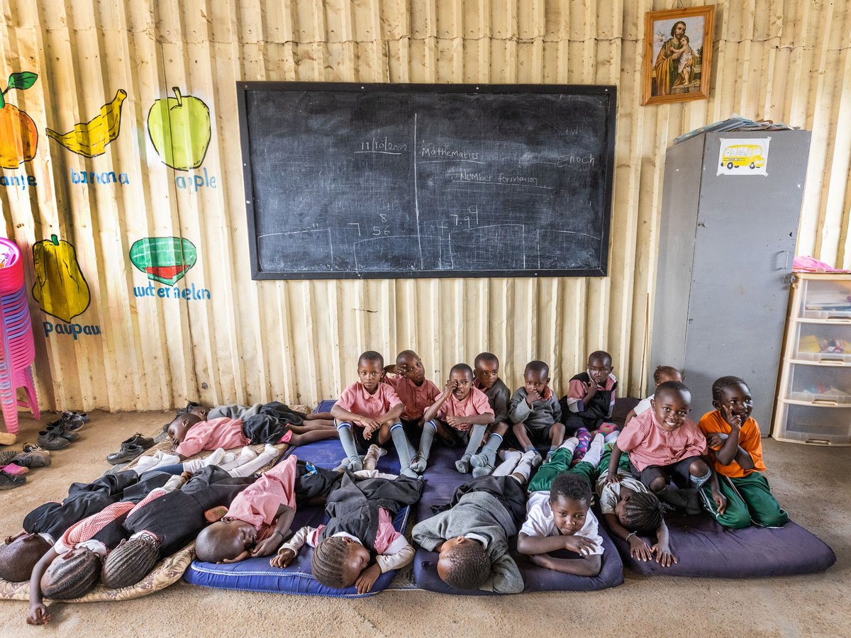 Junge Schüler ruhen und sitzen auf Matten in einem hellen, einfachen Klassenzimmer mit Tafel, Obstzeichnungen und gestapelten Stühlen.