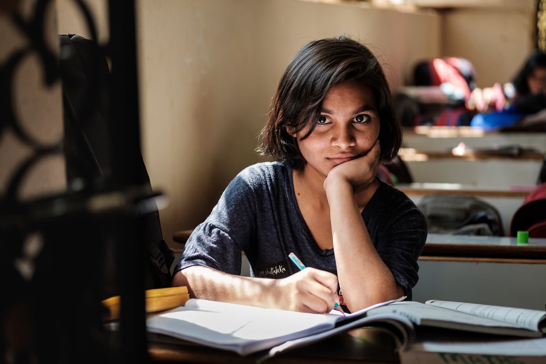Mädchen mit dunklen Haaren schreibt in ein Buch am Schreibtisch und blickt in die Kamera.