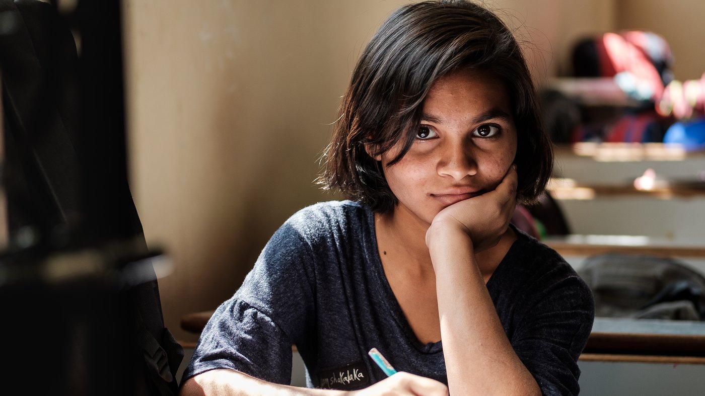Mädchen mit dunklen Haaren schreibt in ein Buch am Schreibtisch und blickt in die Kamera.