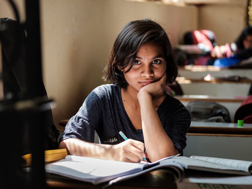Mädchen mit dunklen Haaren schreibt in ein Buch am Schreibtisch und blickt in die Kamera.