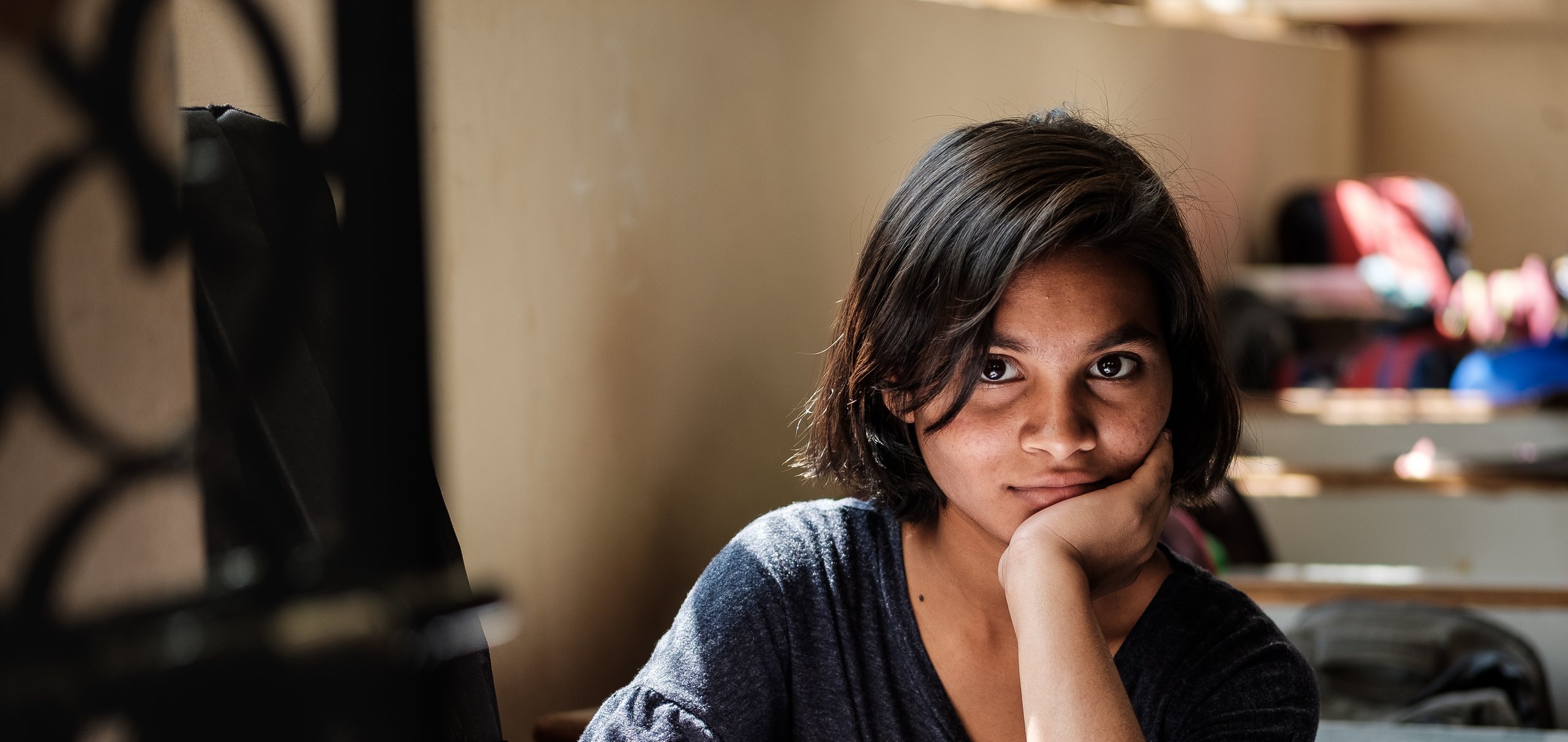 Mädchen mit dunklen Haaren schreibt in ein Buch am Schreibtisch und blickt in die Kamera.