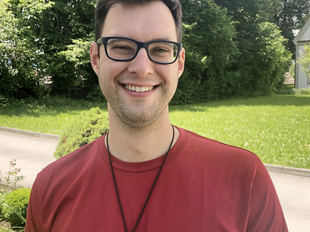 Lächelnder Mann mit Brille und rotem T-Shirt mit einem goldenen Medaillon, draußen vor grünen Bäumen stehend.