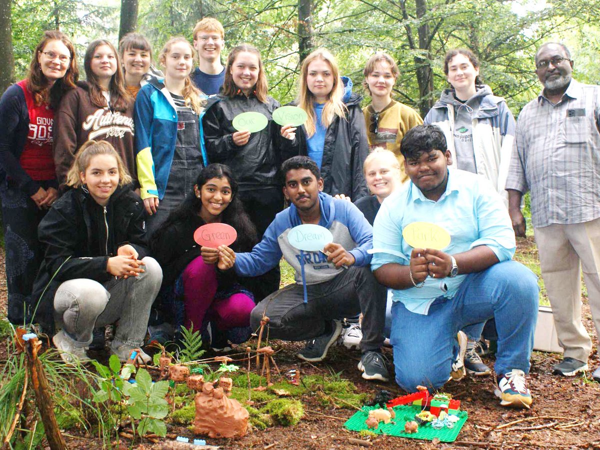 Jugendliche und Erwachsene im Wald mit Schildern ('Green', 'Dream', 'Park') und Naturmodellen auf dem Boden.