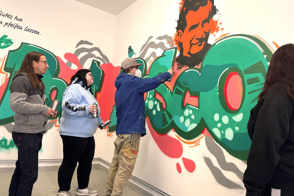 Gruppe von Personen malt gemeinsam ein farbenfrohes Graffiti-Wandbild auf einer weißen Wand, mit Porträt und abstrakten Formen.